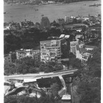 A view from half way up The Peak with a tram ascending the tramway, below is Victoria with to the left center across from the Star Ferry terminal the Hong Kong & Shanghai Bank building.To its right is the open space of the Hong Kong Cricket Club grounds and away further right Causeway Bay and Wanchai. To the right half way down was the Governor's residence. Across the harbor is Kowloon and beyond that the New Territories.