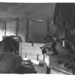 The interior of our tent of our tent on a wet day. The only “furniture” we had were wooden boxes that contain a soldier's gear, to us a "boxes soldier"; you can see an example center above, along with our hanging uniforms. The tents were difficult to keep tidy in those conditions, and there was not a great deal of comfort. The object in the middle rear is Ian Styles collapsed on his bed.