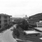 Here is a general view of Stanley Barracks to the south looking toward the tip of the peninsula and taken from the veranda of our barrack block. On left is the barrack block occupied by X Troop and BHQ, then the Sergeant's Mess and the Garrison Church of St Barbara. To the right is the cookhouse and below that the ablutions, lower down is the vehicle park and Square. The figures in the road are Ian Styles, Jim Dallaway and McKenny who are going to fetch the dhobi from the building to the far right.