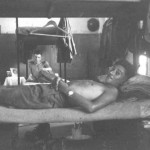 Jock Lyon, in front, is prone on my bunk. Behind him is Ian Styles on his. To the right is my boxes soldier, below are my plate, mug and eating irons next my boots. Above my bunk are uniforms on hangers. The shiny object on the wall behind Ian is his steel helmet. To the left is the open fireplace. A domestic note is the orange box next to Ian with some personal photographs on it belonging to the occupant of the upper bunk. Note the lack of comforts; e.g., a decent mattress, no sheets for the bed (though I think we had them), and no furniture, along with the bare barrack room.